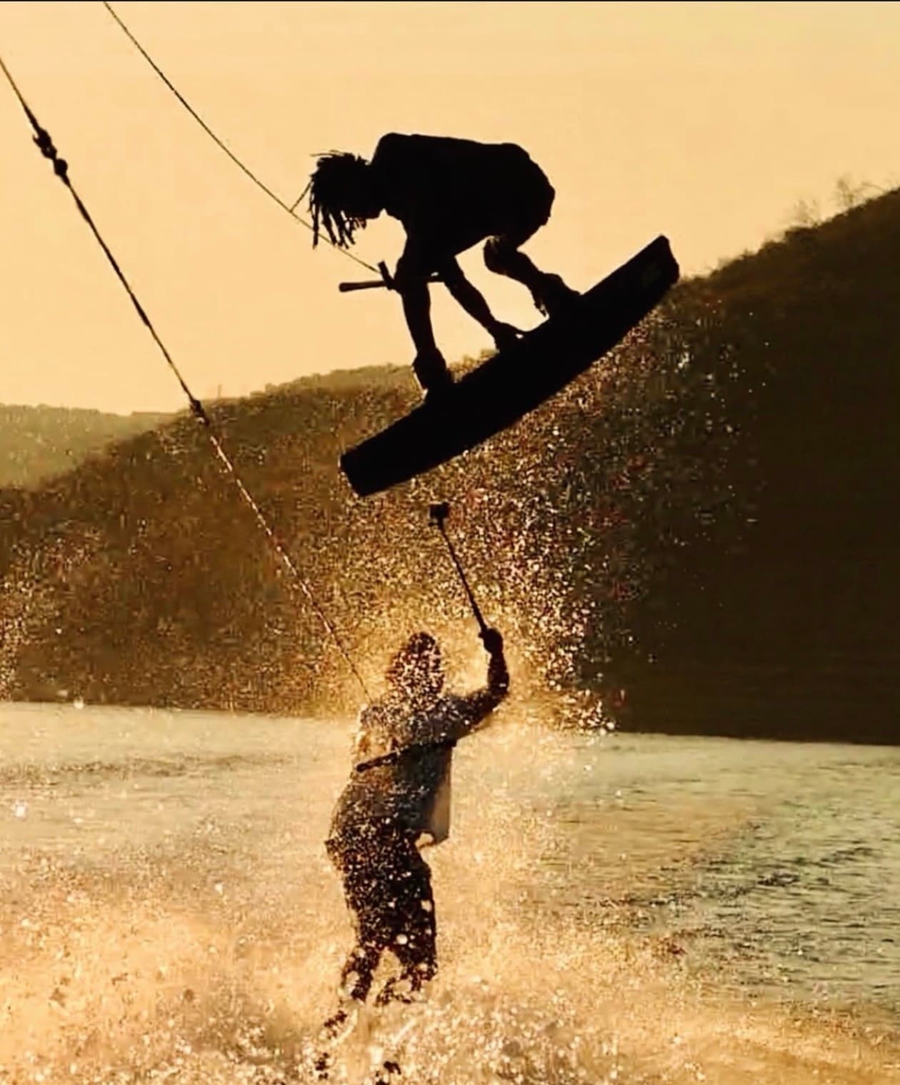 A wakeboarder silhouetted against golden spray above another rider